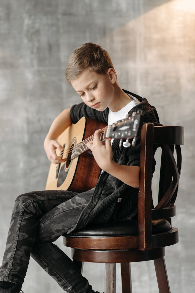 A boy sitting on a chair playing an acoustic guitar indoors, focusing intently.